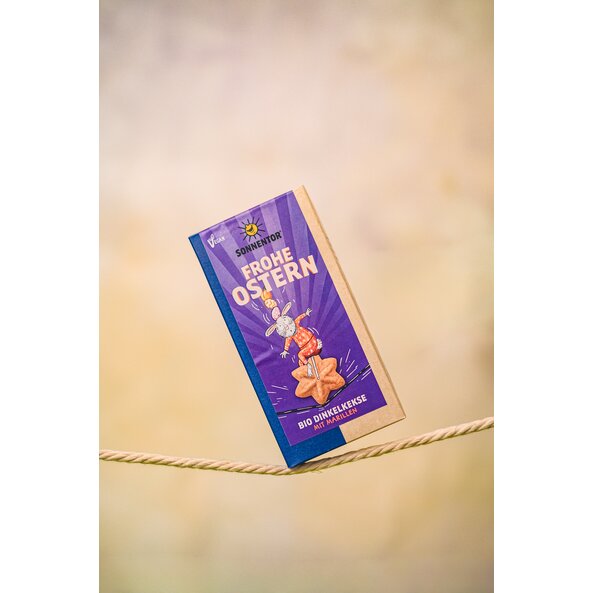 The Easter biscuits are balancing on a rope. On the biscuit label, the Easter Bunny is riding an Easter biscuit like a circus performer on a unicycle on a rope, balancing Easter eggs and an apricot on his head. 